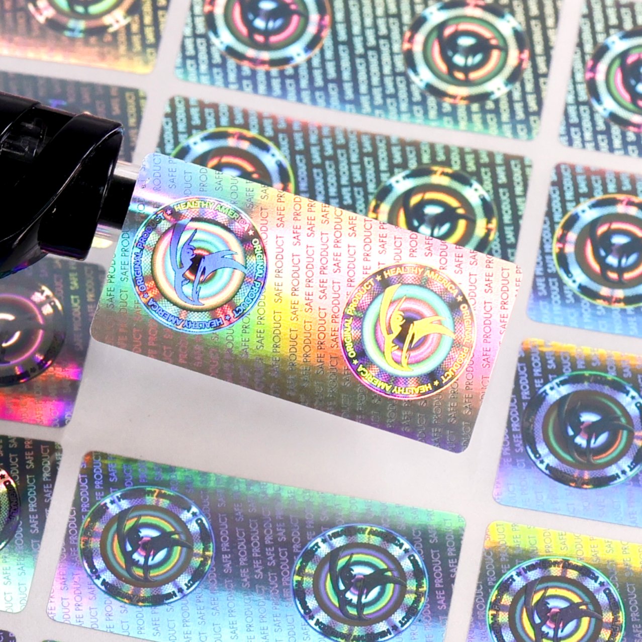 Production sheet of laser-engraved holographic labels — client logo surrounded by SAFE PRODUCT microtext, printed full-sheet then die-cut