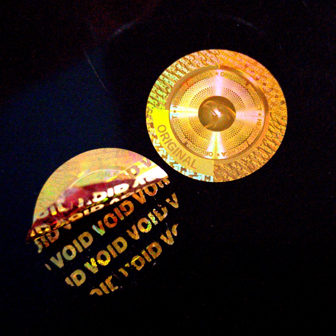 Gold laser-engraved ORIGINAL holographic label — intact label showing ORIGINAL text, and a peeled one revealing VOID pattern on the black surface