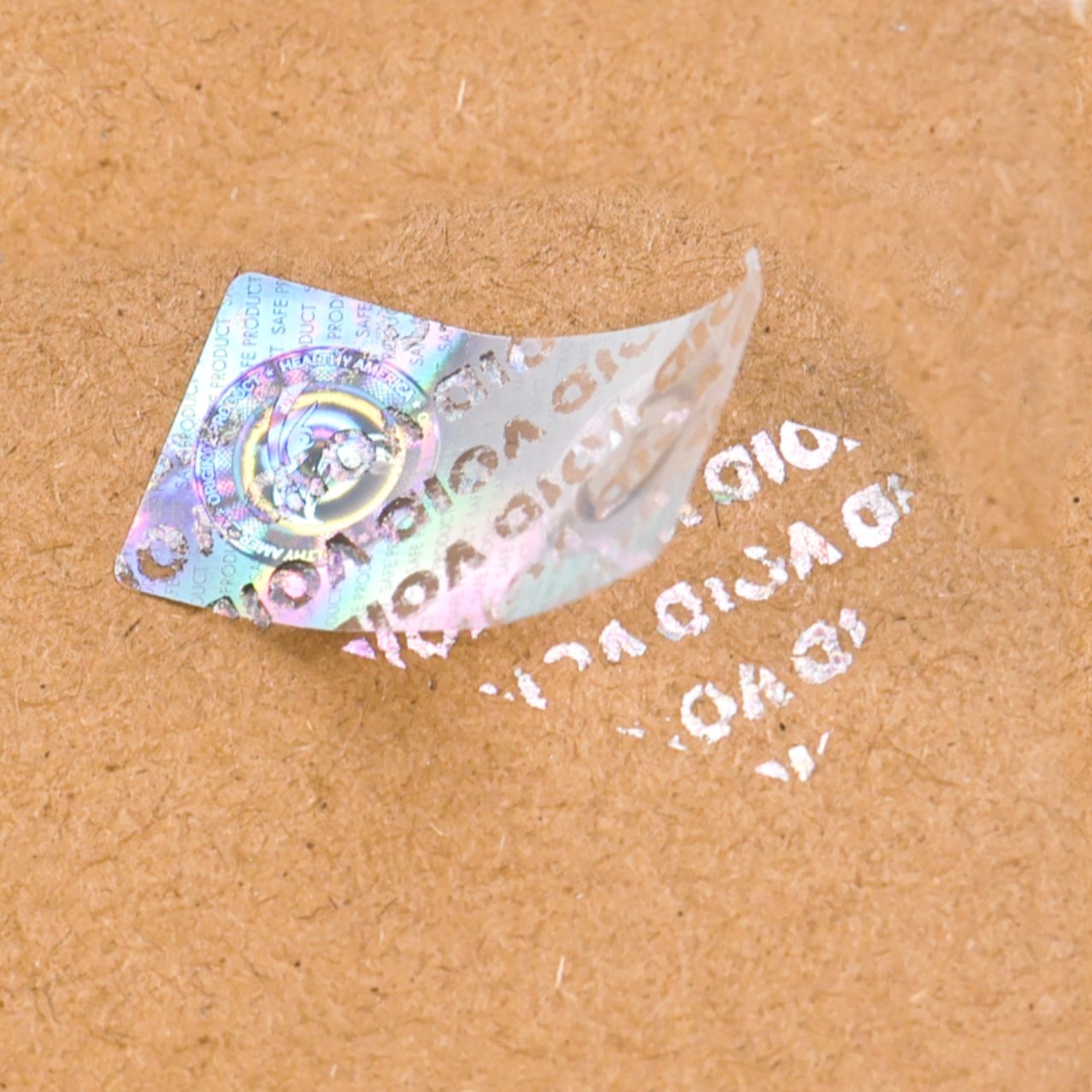 Silver laser-engraved holographic label with SAFE PRODUCT microtext being peeled on kraft paper, VOID pattern left behind